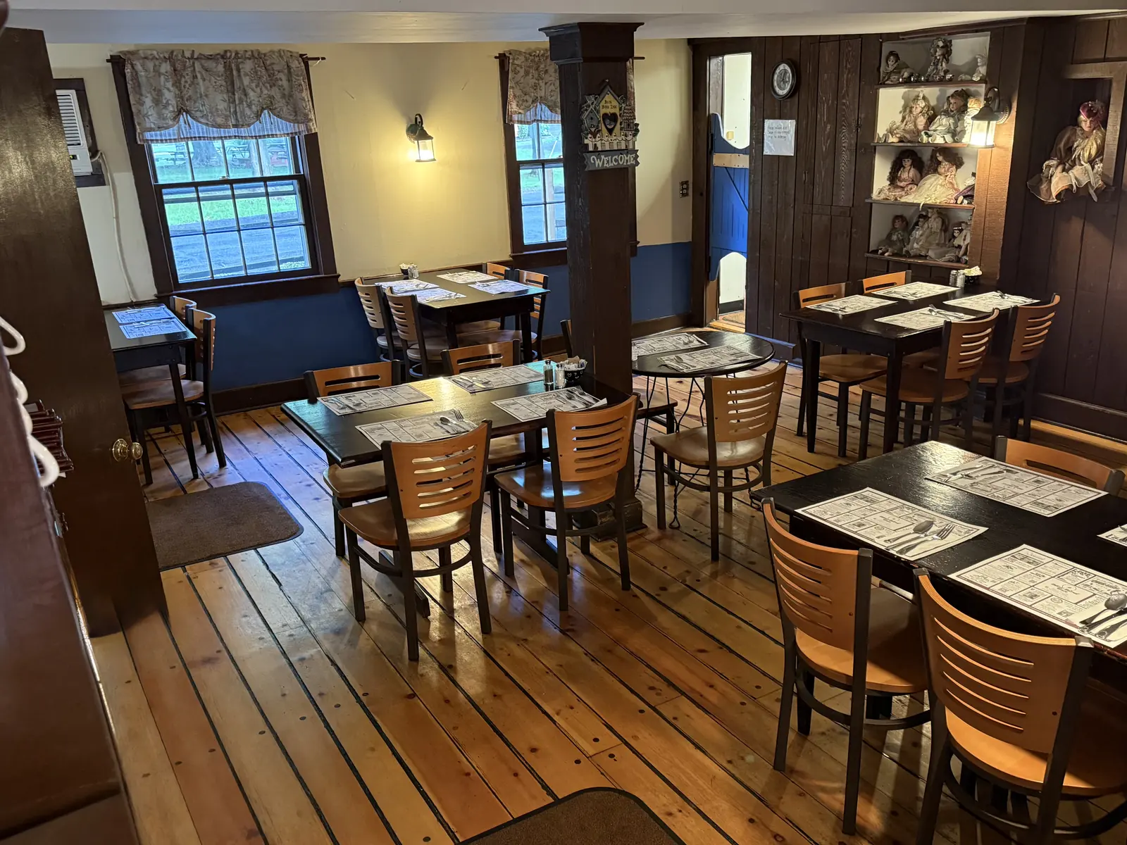 The empty dining room: wide plank floors, blue wainscoting, tables set with paper placemats, two windows onto the road, a Welcome sign on the centre post and shelves of porcelain dolls along the back wall